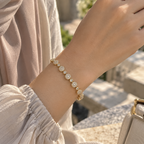 A delicate gold bracelet with round floral clusters of clear stones, shown worn on a person's wrist. The person is wearing a light beige sleeveless top, and the background is a softly blurred outdoor stone setting. Celestial Rhythm Zirconia Bracelet 18k gold plated harma jewelry