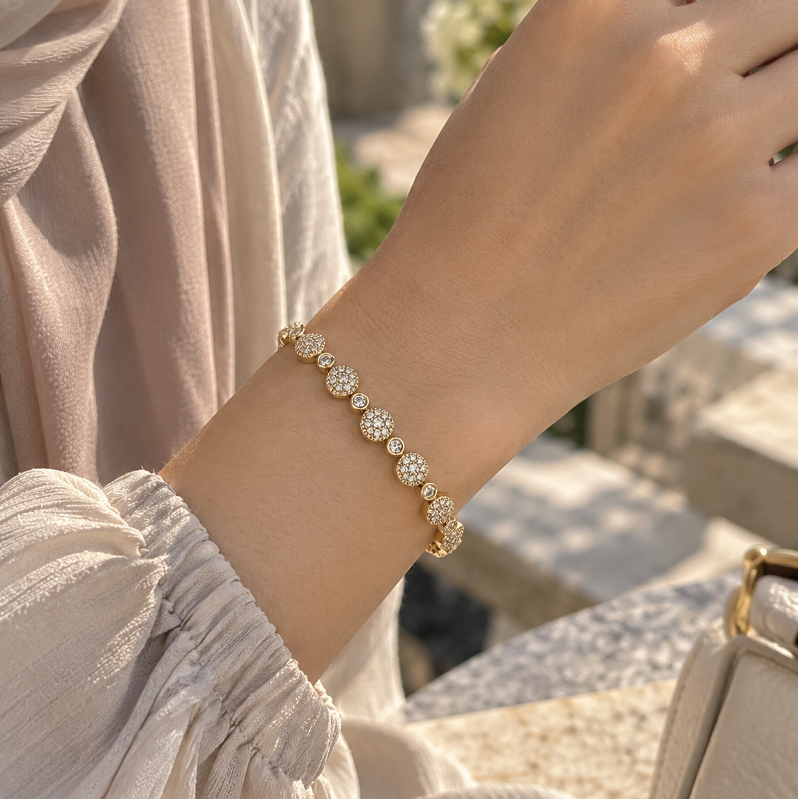 A delicate gold bracelet with round floral clusters of clear stones, shown worn on a person's wrist. The person is wearing a light beige sleeveless top, and the background is a softly blurred outdoor stone setting. Celestial Rhythm Zirconia Bracelet 18k gold plated harma jewelry