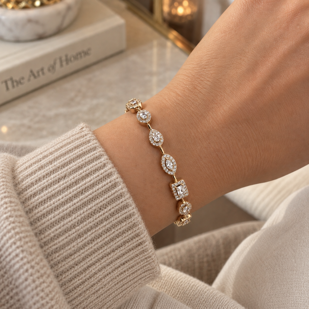 A gold bracelet with multiple oval and square links set with sparkling clear stones, shown worn on a person's wrist. The person is wearing a beige ribbed knit sweater, with a blurred background featuring a book and warm indoor lighting. Fall Constellation Zirconia Bracelet 18k gold plated Harma jewelry 3 