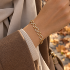 A gold chain link bracelet with small clear stones, worn on a woman's wrist. The image shows a close-up of her hand, sleeve, and scarf, with a blurred background of autumn leaves. Gilded Spark Gold Zirconia Link Bracelet | Harma Jewelry 18k gold plated