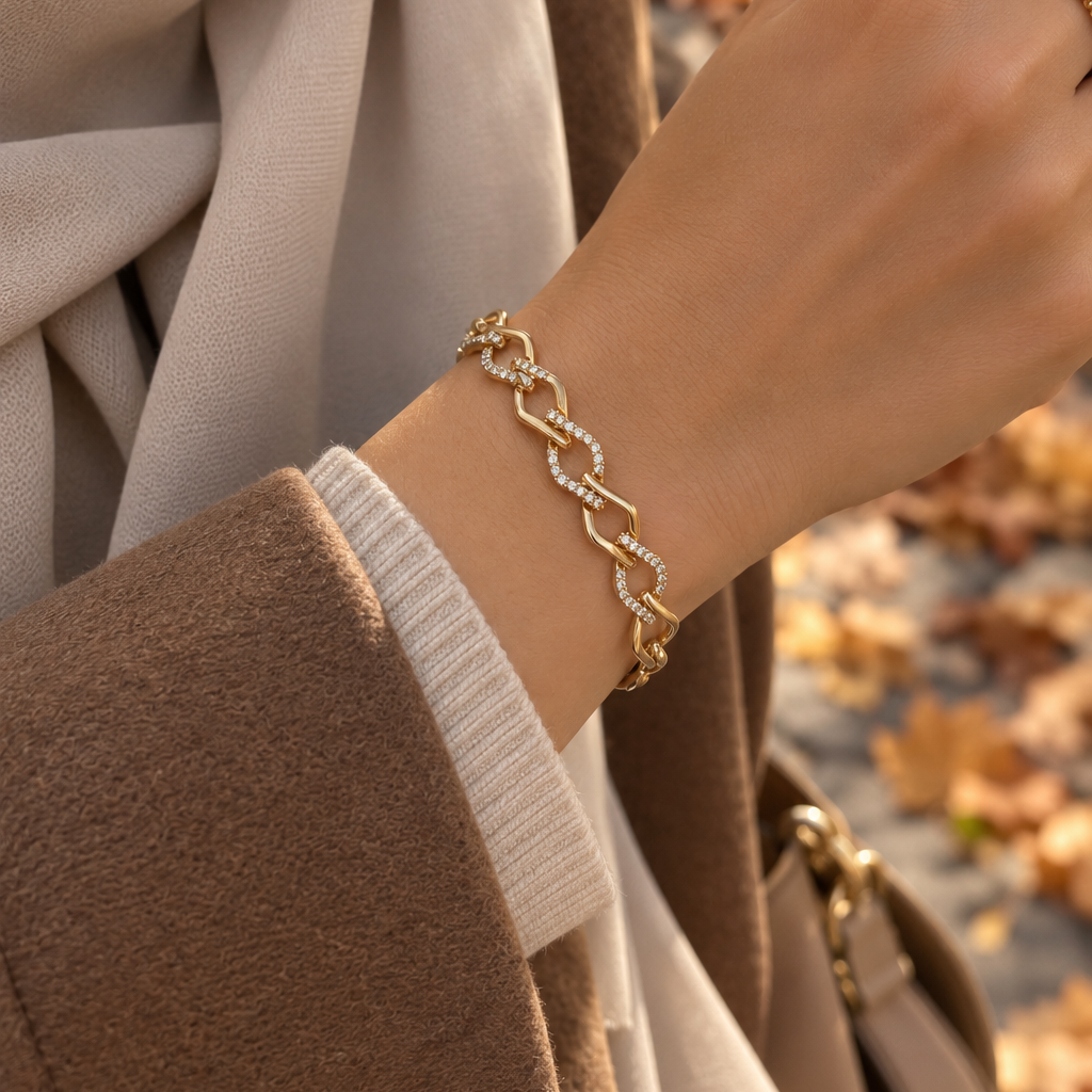A gold chain link bracelet with small clear stones, worn on a woman's wrist. The image shows a close-up of her hand, sleeve, and scarf, with a blurred background of autumn leaves. Gilded Spark Gold Zirconia Link Bracelet | Harma Jewelry 18k gold plated