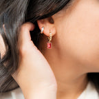 Close-up of a person wearing gold earrings with red gemstones, holding their hair back. Gold ring with red gemstones on a white background. Harma Gold plated Rosy Revival Link Chain Crystal Hoop 