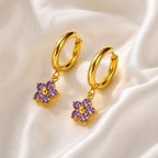 Gold hoop earrings with purple flower-shaped charms on a satin surface