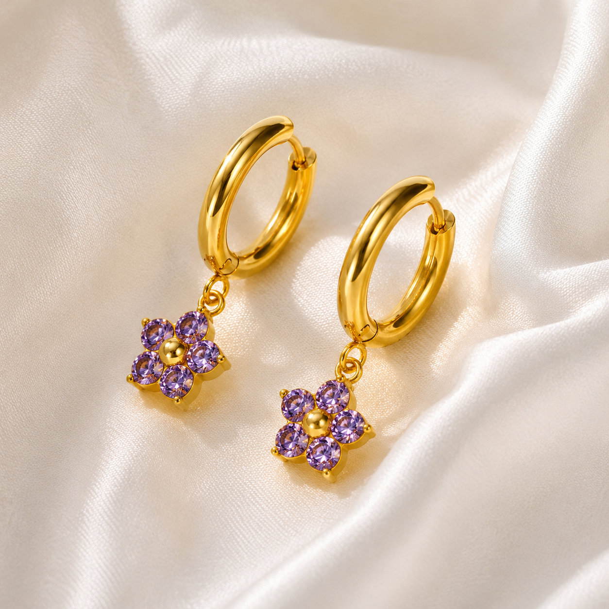 Gold hoop earrings with purple flower-shaped charms on a satin surface