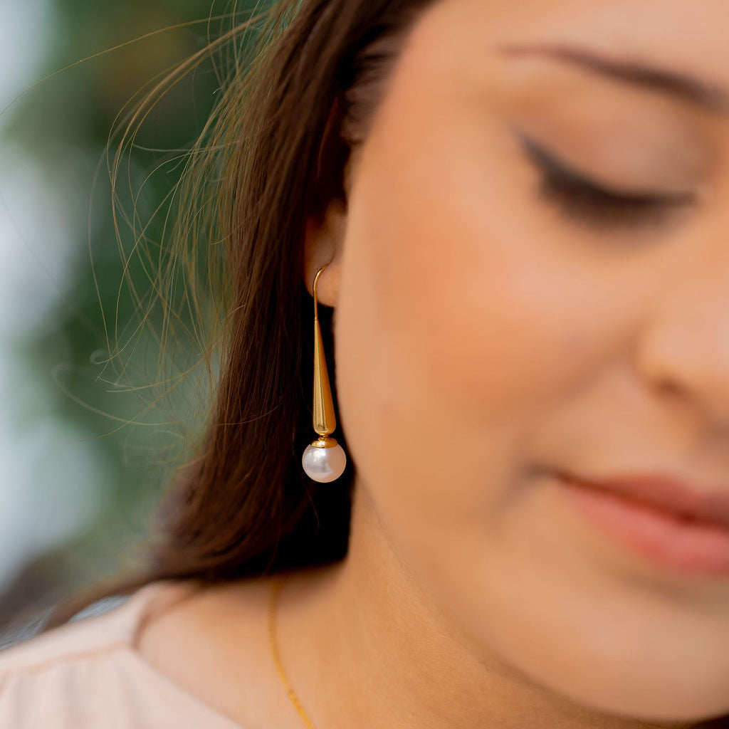 Close-up of a woman wearing gold earrings and a necklace with a blurred background. Harma Gold Plated Pearl I Am Grounded Dangle Earrings