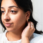 Woman wearing gold earrings and a ring, touching her ear against a plain background. Harma Gold Plated Resilient Spark Gold Earrings