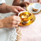Two people holding gold teacups on a decorative table. With bold princess cut Zirconia ring 