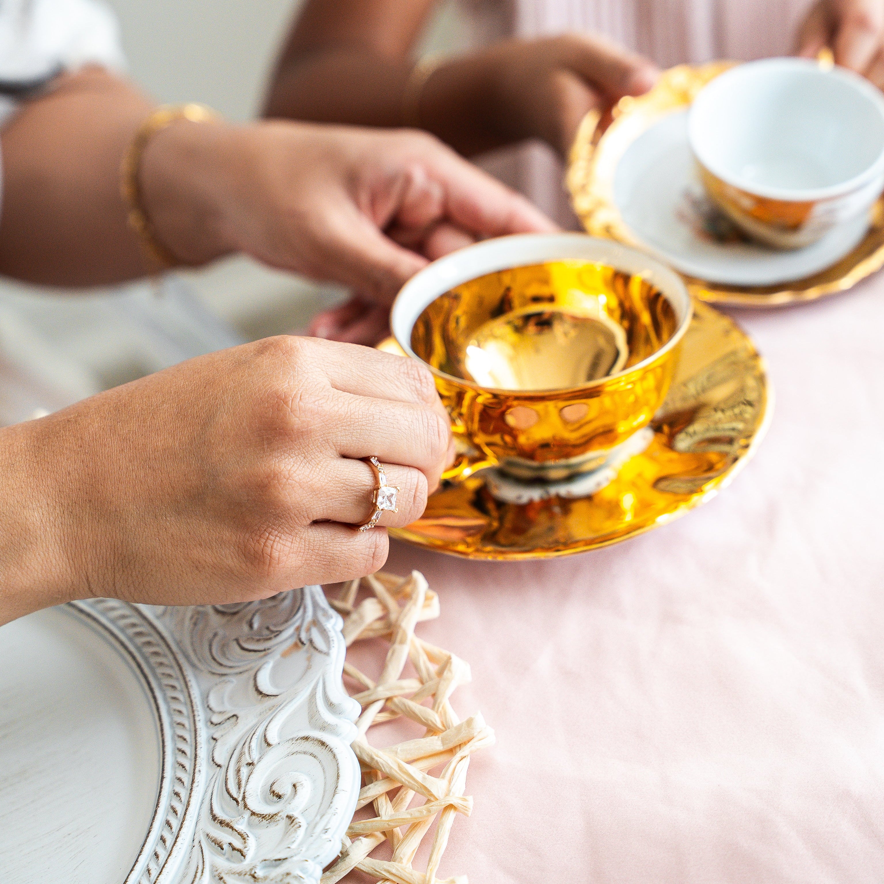 Two people holding gold teacups on a decorative table. With bold princess cut Zirconia ring 
