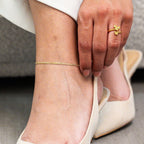 Close-up of a person's foot wearing a gold anklet and gold ring on a neutral background