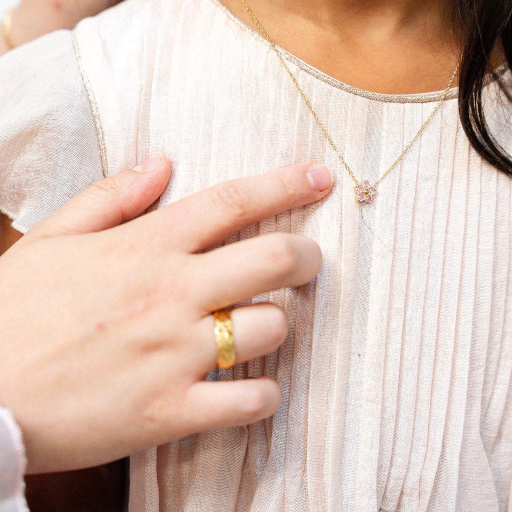 Person wearing a gold necklace with a small pendant, held by another person. Harma_Jewelry_Gold_Plated_Little_Bloom_PINK_Zirconia_Necklace_2