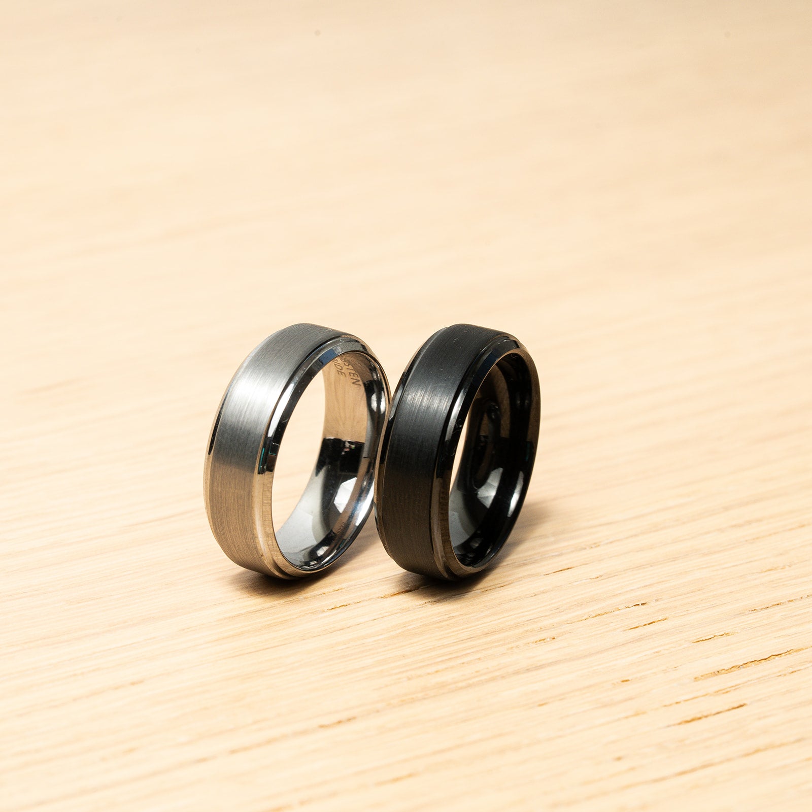 Two rings, one silver and one black, on a wooden surface Harma Jewelry Men Tungsten Carbide Band Ring Black Silver Simple 