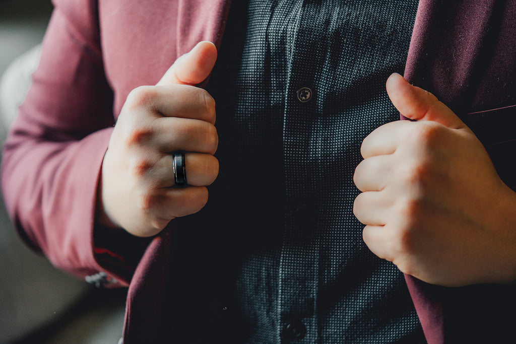 Person wearing a dark shirt with a pink jacket, adjusting the jacket with both hands. Harma Jewelry Men Tungsten Carbide Band Ring Black Simple 