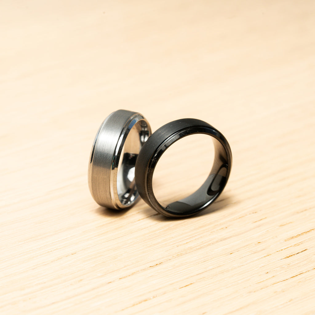 Two rings, one silver and one black, on a wooden surface. Harma Jewelry Men Tungsten Carbide Band Ring Silver Black Simple 
