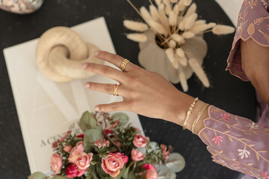 Hand with gold rings on a table with flowers and a book Harma Luminous Noor Lantern Bracelet