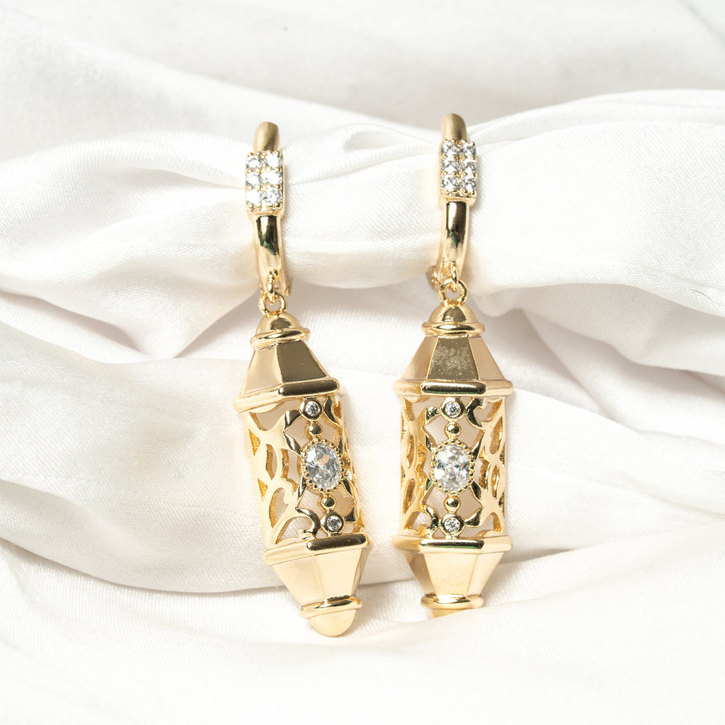 Gold earrings with diamond accents on a white satin background. Harma Luminous Noor Lantern Necklace Set