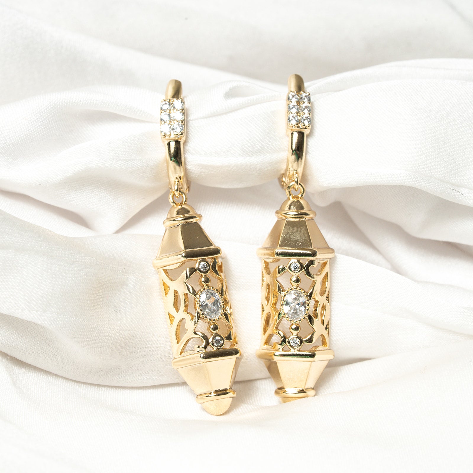 Gold earrings with diamond accents on a white satin background. Harma Luminous Noor Lantern Necklace Set