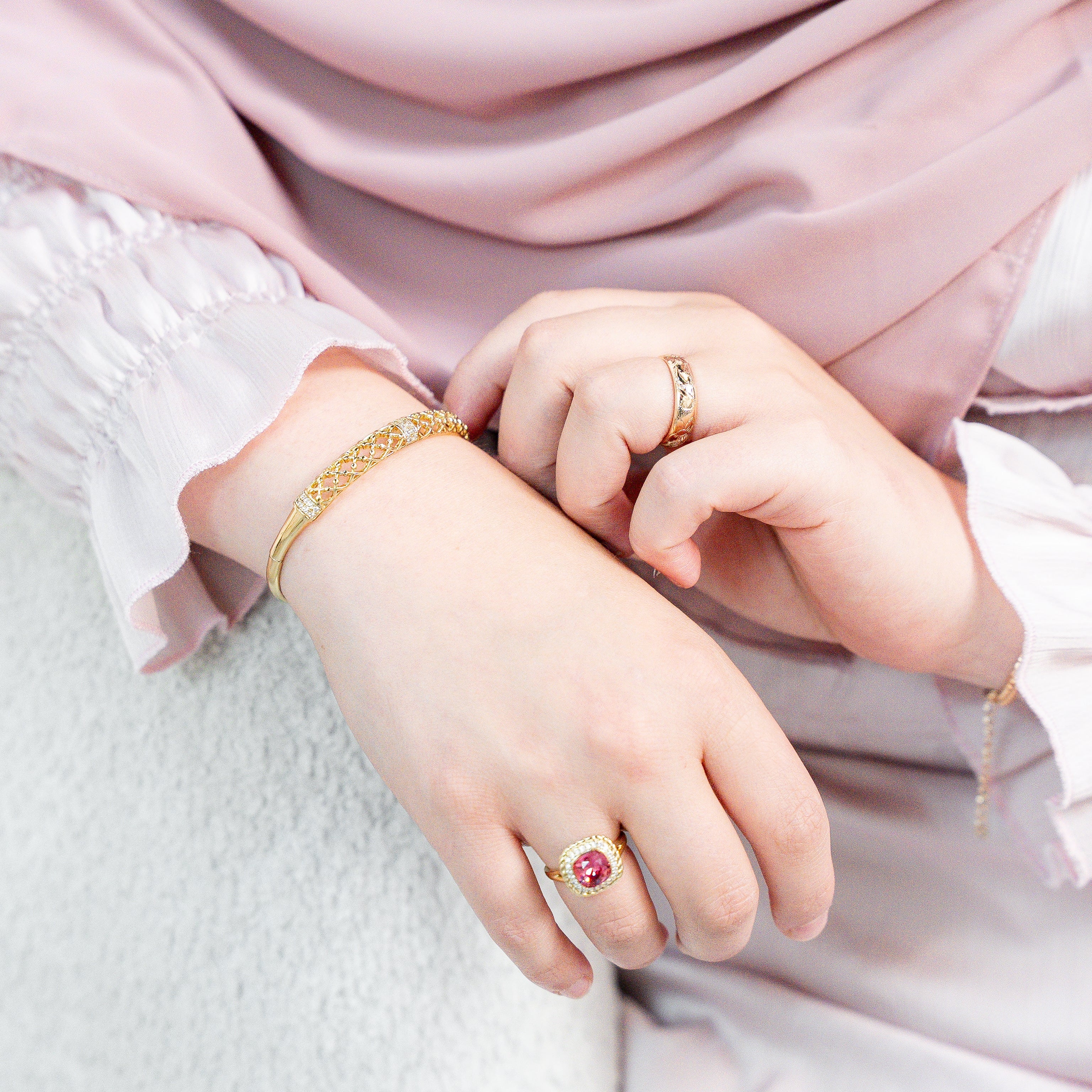Close-up of hands wearing gold bracelets and rings with a soft pink fabric background. Harma jewelry gold plated Golden Heritage Bangle Bracelet