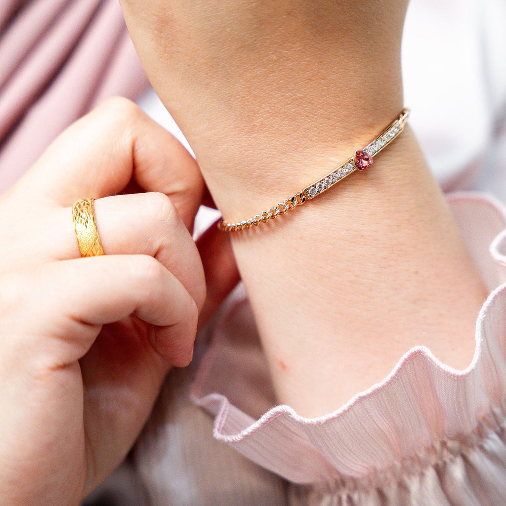 Close-up of a child's hand wearing a gold ring and bracelet on a pink fabric background. Harma jewelry gold plated I Am Loved Crystal Heart Bracelet