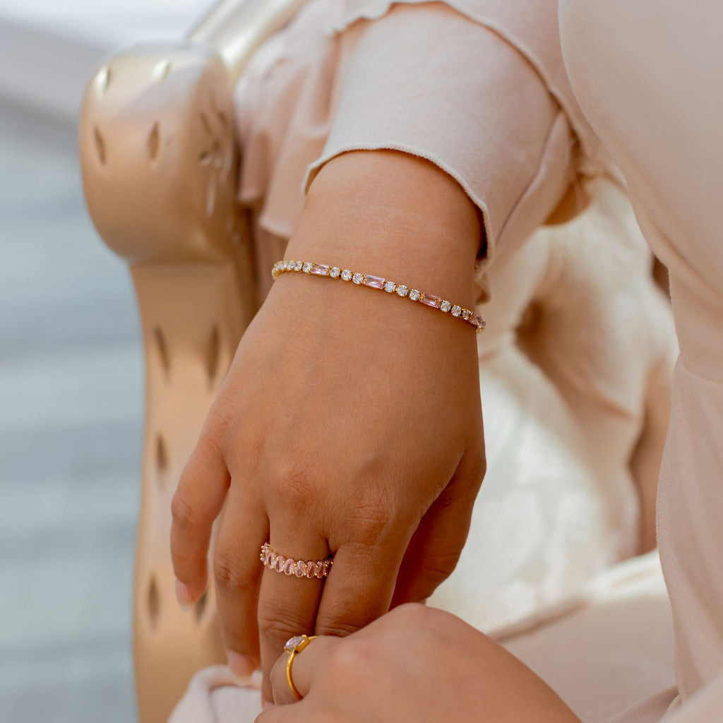 Close-up of hands wearing rings and bracelets with a blurred background. Harma jewelry gold plated Moments of PINK Tennis Bracelet