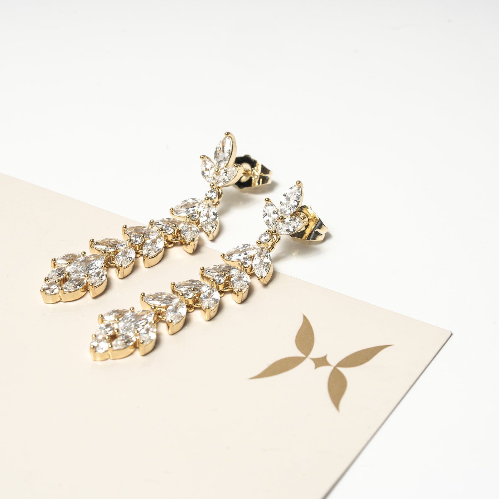 Harma jewelry gold plated chandelier earrings zircnoia hypoallergenic 14k. Gold earrings with clear stones on a beige card with a logo