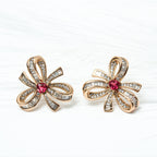 Pair of gold flower-shaped earrings with red gemstones on a white background. Harma gold plated I Am Radiant Bow Stud Earrings