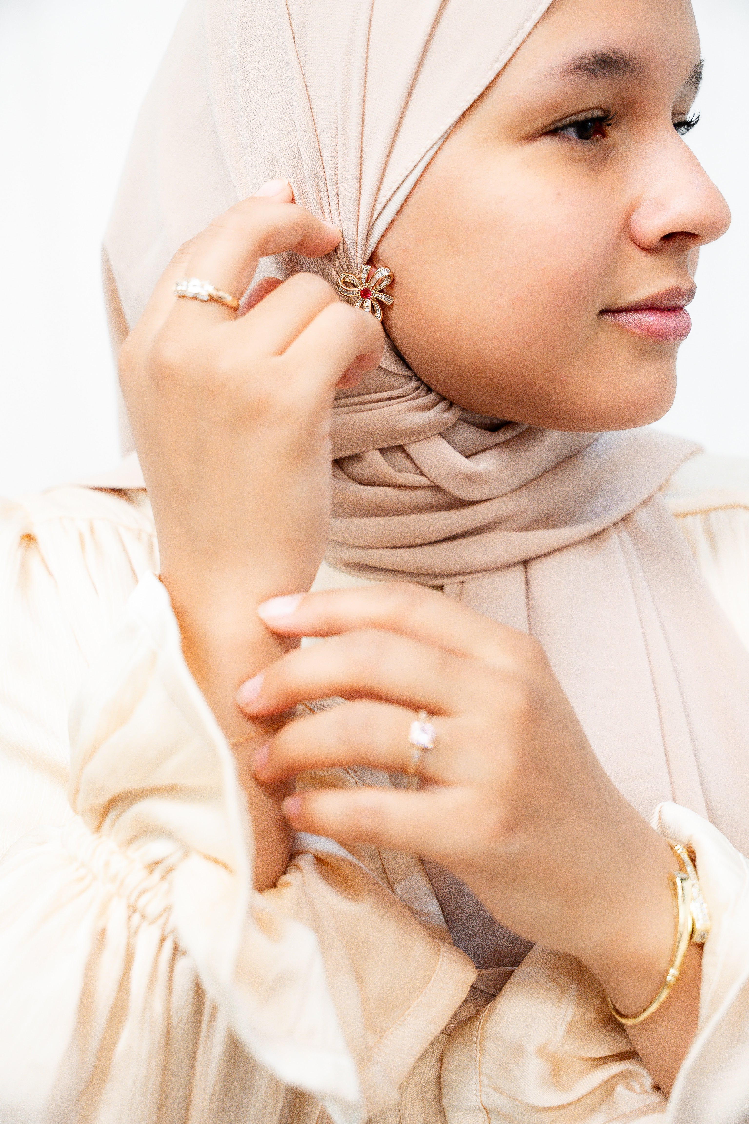 Woman adjusting a headscarf with a close-up of her hands and face. Harma gold plated I Am Radiant Bow Stud Earrings