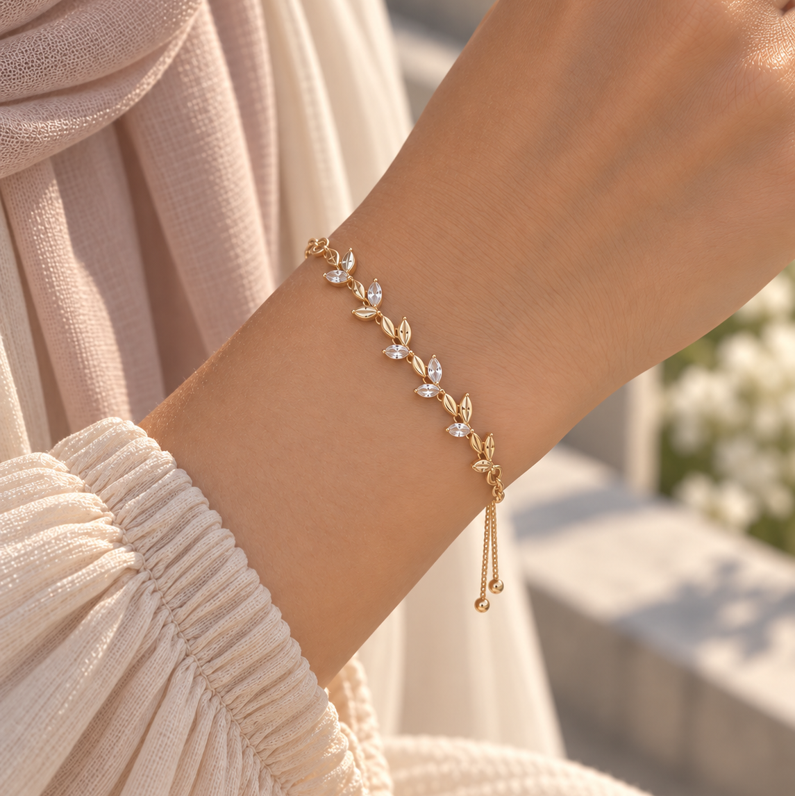A delicate gold leaf bracelet with clear crystal accents worn on a person's wrist, paired with a cream-colored pleated top and a soft beige scarf. Whispering Magnolia Marquise Adjustable Bracelet 18k gold plated Harma Jewelry