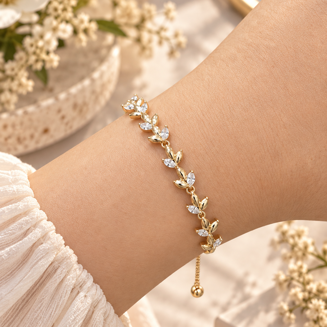 A delicate gold bracelet with leaf and flower motifs set with clear crystals, worn on a woman's wrist. The bracelet features a small dangling gold bead at the end. Whispering Magnolia Marquise Adjustable Bracelet 18k gold plated Harma Jewelry