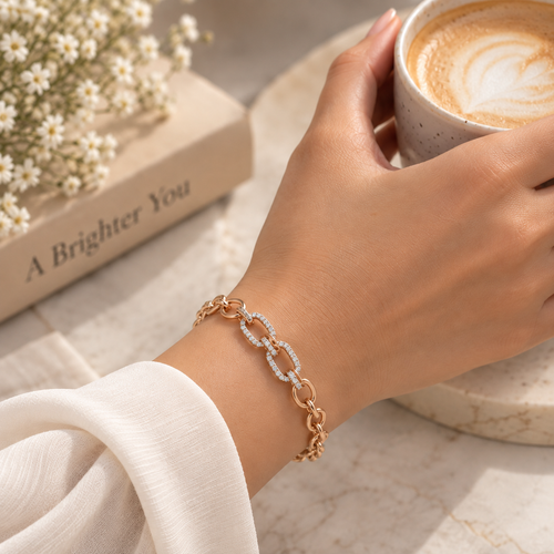 A close-up lifestyle shot of a person's hand holding a cup of latte while wearing a delicate rose gold chain bracelet with alternating links, one of which is lined with small clear crystals. The background features a marble surface, a small book with white daisies, and a warm, soft-focus aesthetic. Élan Pavé Link Zirconia Bracelet 18k gold plated harma jewelry