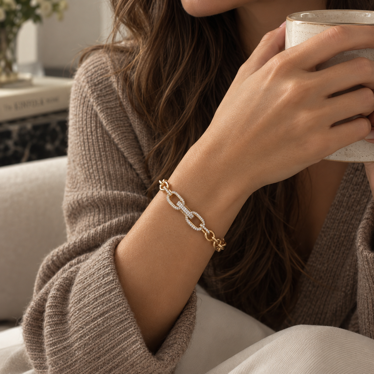 A close-up lifestyle shot of a woman wearing a gold chain-link bracelet with pavé-set clear stones on her wrist, holding a ceramic mug while wearing a brown knit cardigan. Élan Pavé Link Zirconia Bracelet 18k gold plated harma jewelry